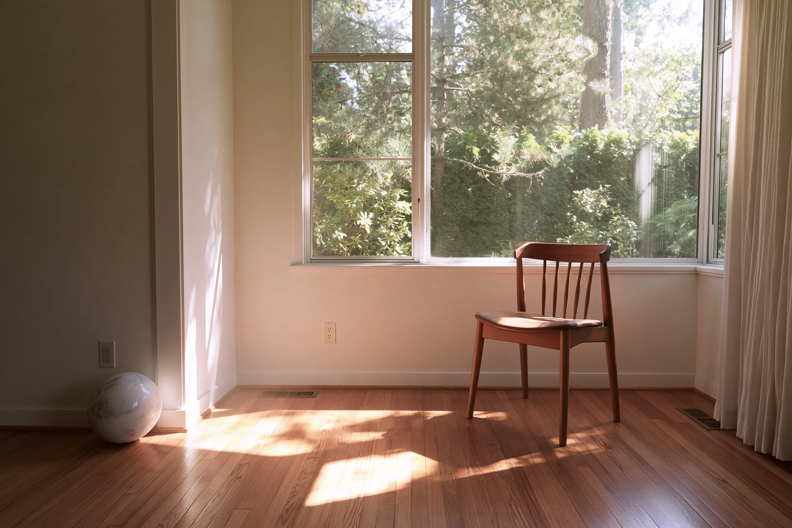 Empty living room of a house with a hardwood floor, a wooden chair by the window and a small ball resting against the wall in the corner
